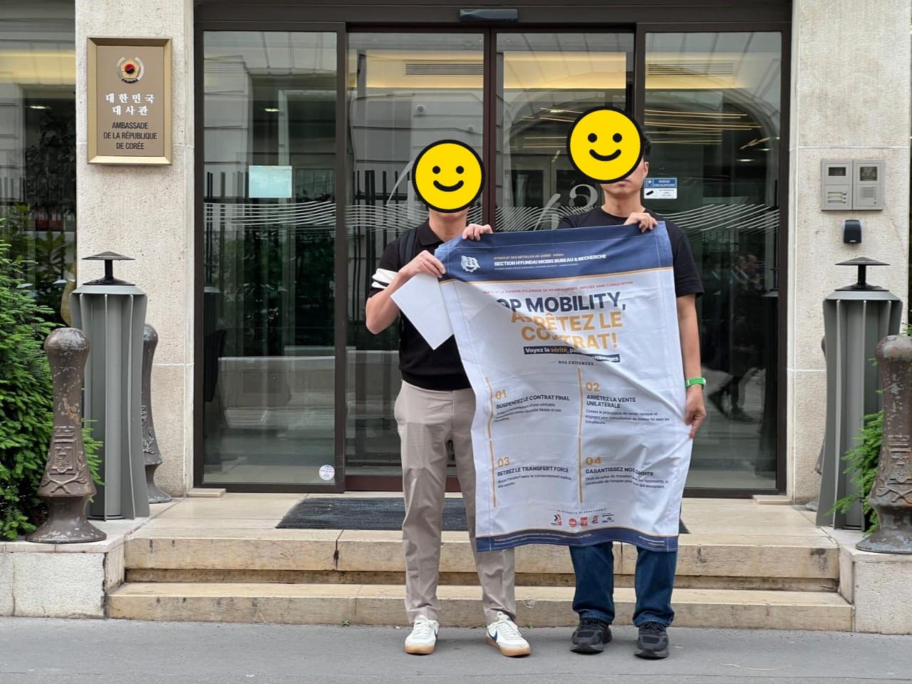 프랑스 주재 대한민국 대사관 명패 옆에서 두 조합원이 'OP MOBILITY, ARRÊTEZ LE CONTRAT!' 프랑스어 현수막을 펼쳐 든 모습