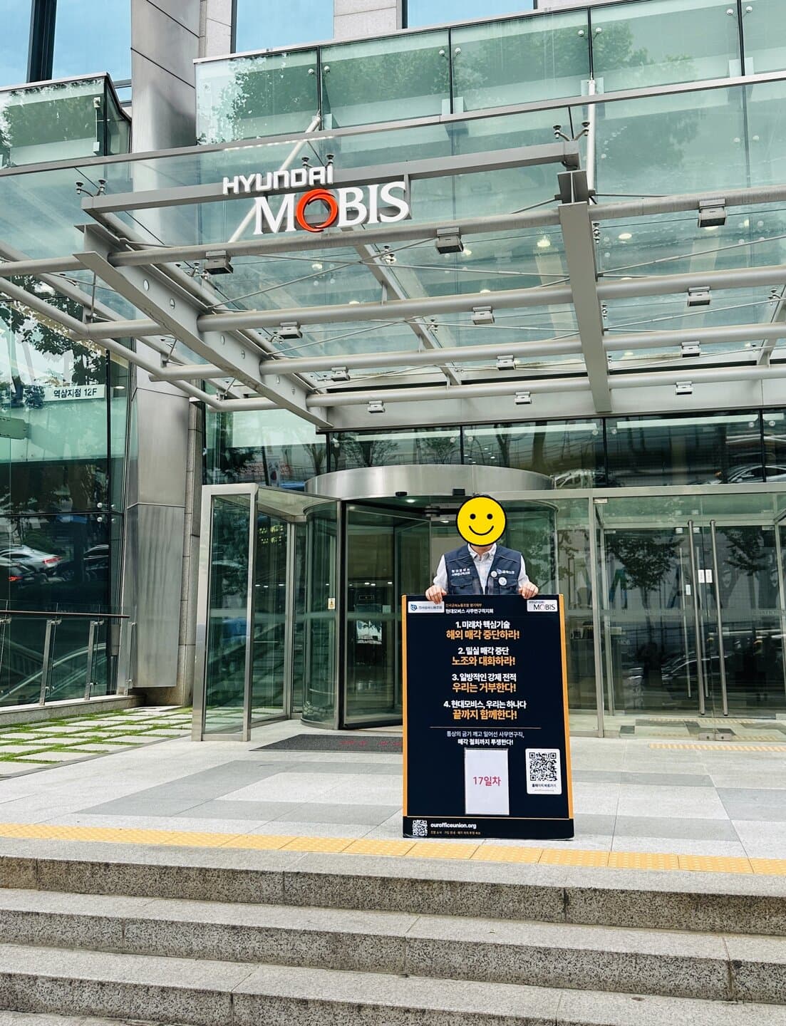 현대모비스 본사 정문 앞, 회전문을 배경으로 네 가지 요구가 적힌 1인시위 폼보드 뒤에 선 조합원이 보이는 17일차 광경