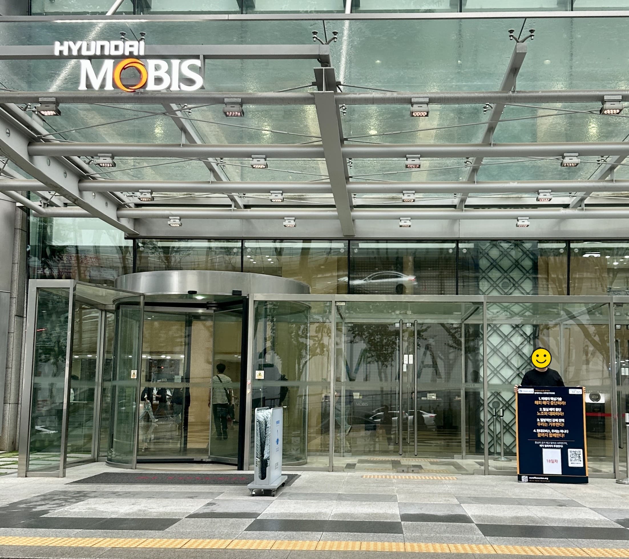 현대모비스 본사 정문 앞 회전문을 배경으로, 네 가지 요구와 18일차 표시가 적힌 1인시위 폼보드 옆에 선 조합원이 보이는 18일차 광경