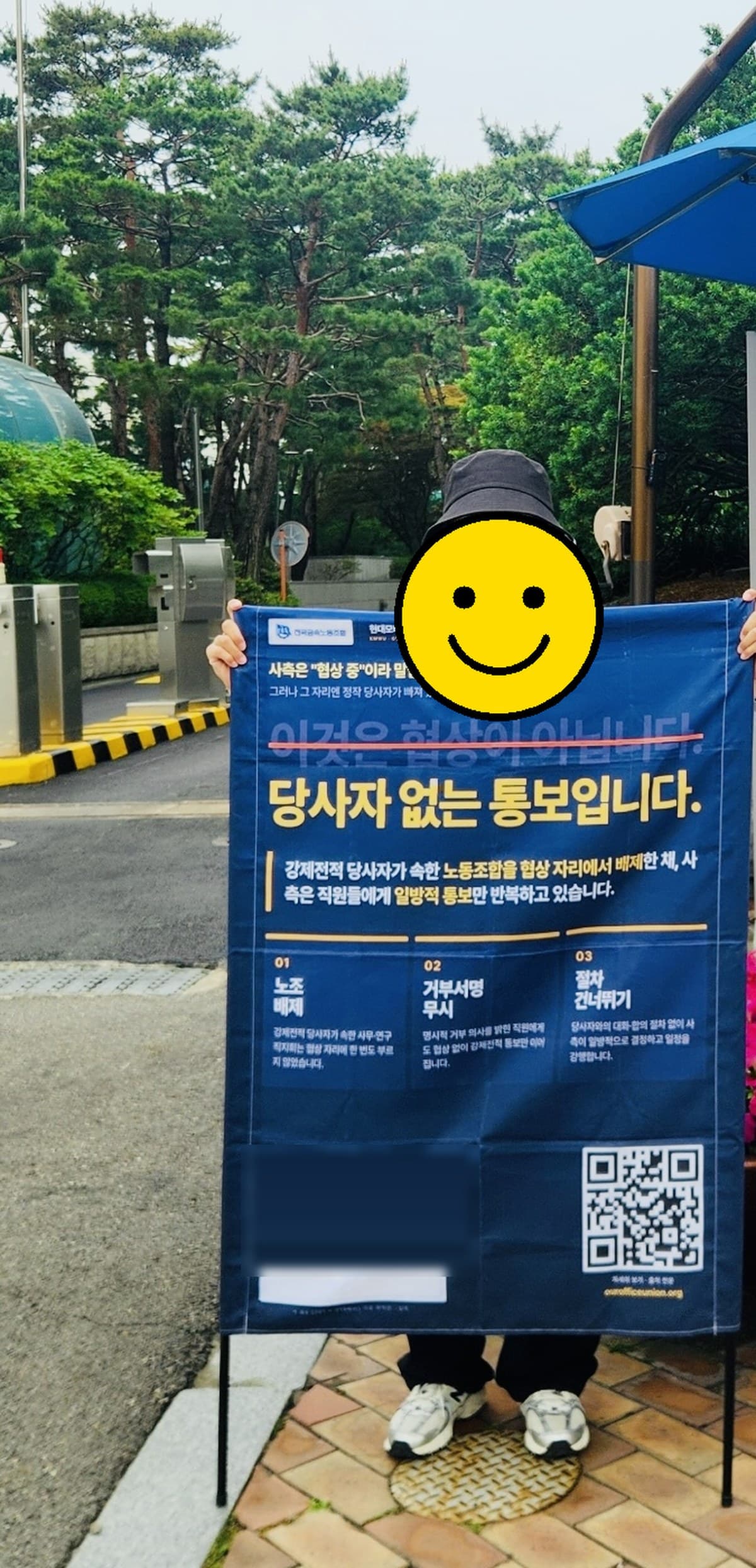 서울 양재동 현대자동차그룹 본사 앞에서 “당사자 없는 통보입니다” 폼보드를 들고 선 현대모비스 사무연구직지회 조합원의 모습 (얼굴은 스마일 이모지로 가림 처리)